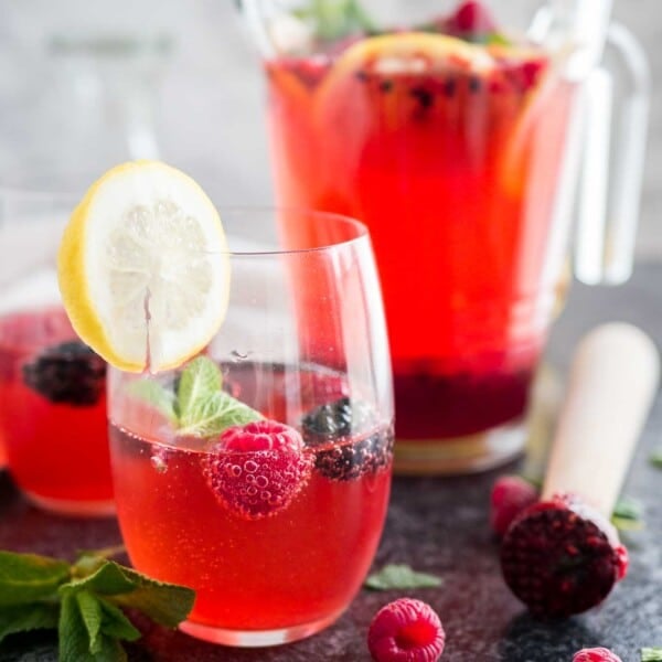 Two glasses of raspberry lemonade with raspberries and mint in them and a slice of lemon on the rim. There's a jug with the rest of the lemonade in the background and a muddler next to it.