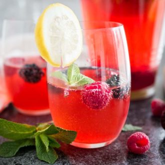 Close-up of two glasses of raspberry lemonade with raspberries and mint in them and a slice of lemon on the rim. There's a jug with the rest of the lemonade in the background.
