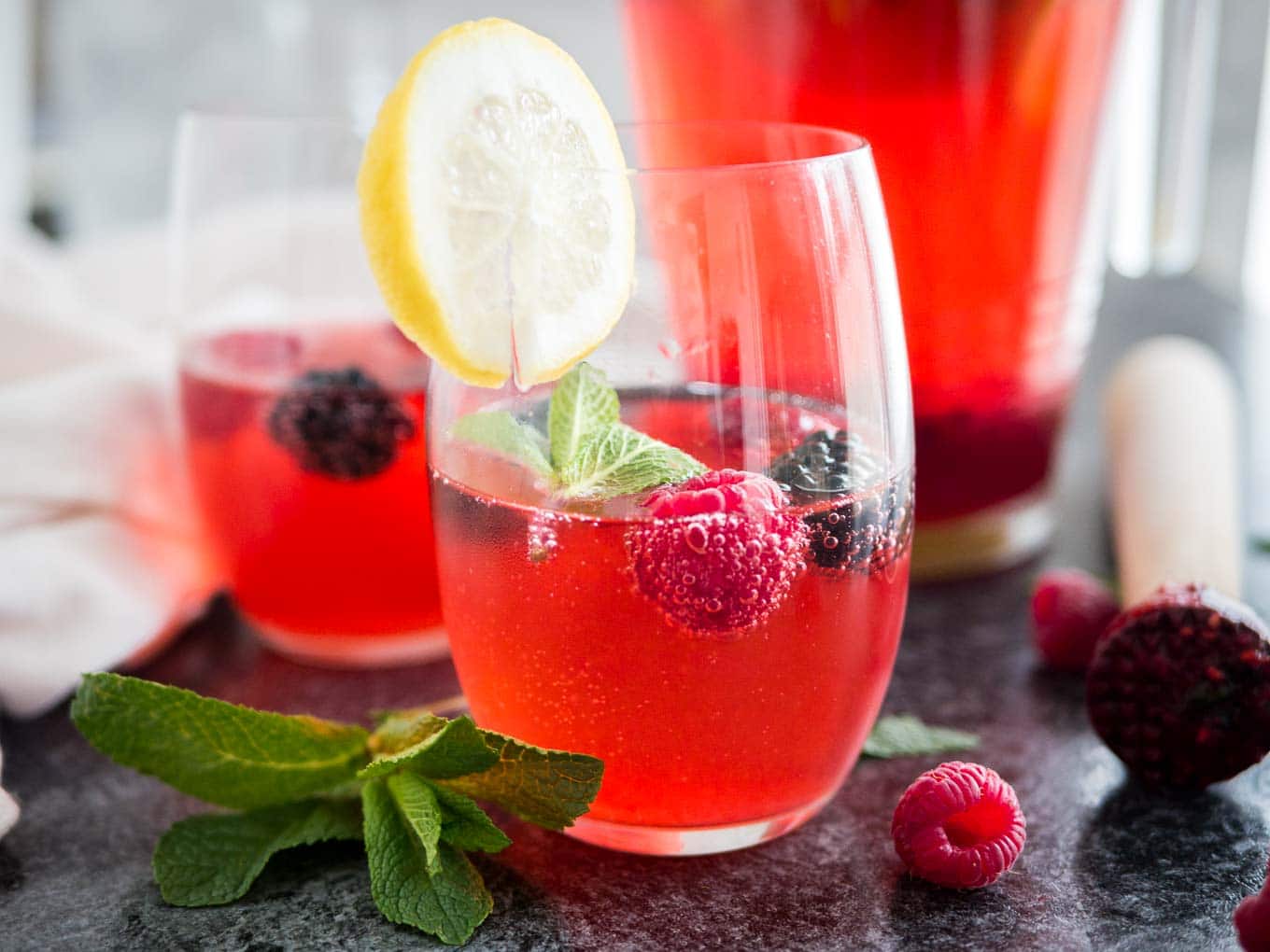 Close-up of two glasses of raspberry lemonade with raspberries and mint in them and a slice of lemon on the rim. There\'s a jug with the rest of the lemonade in the background.