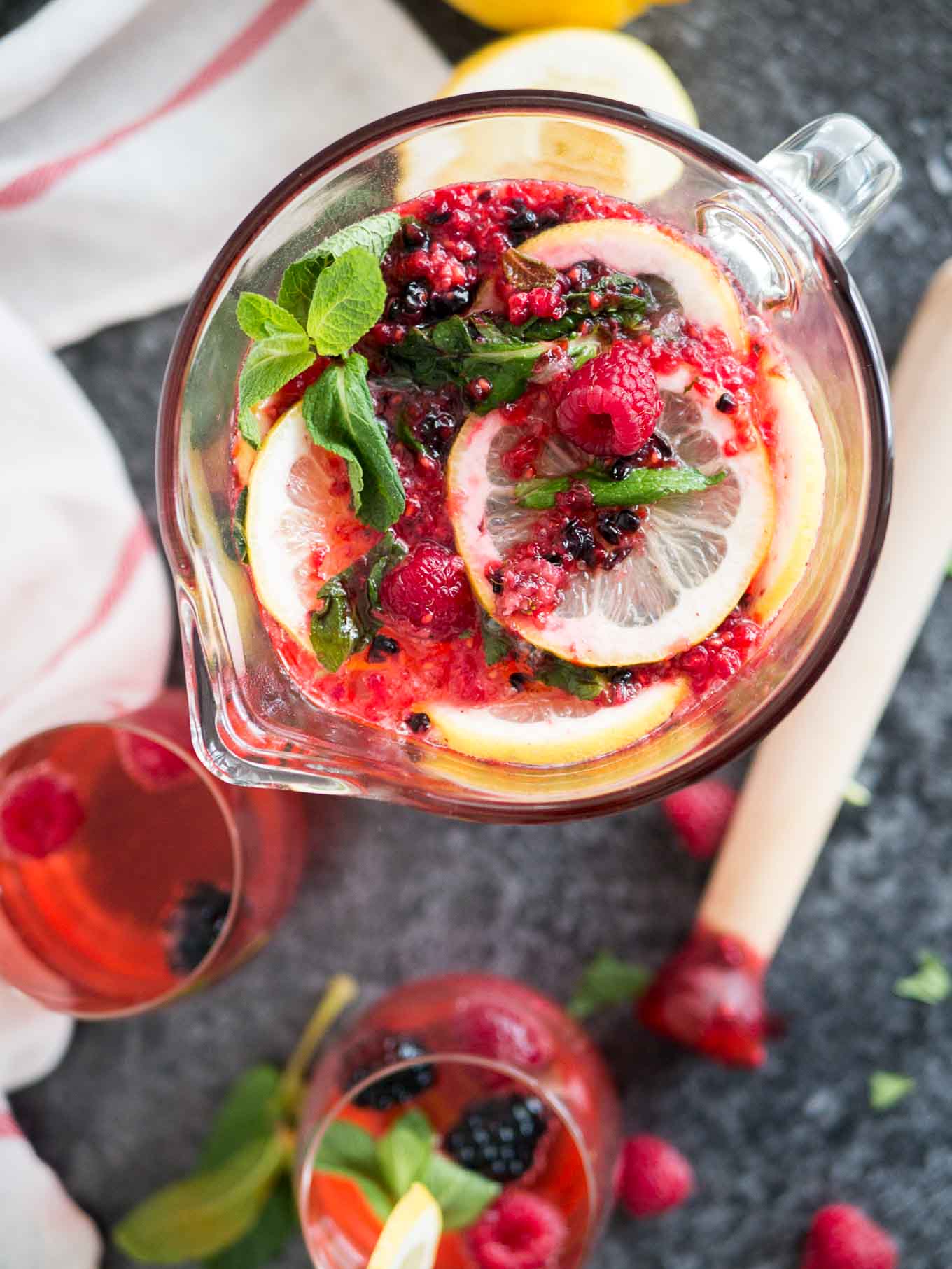 Top-down shot of a glass pitcher with homemade raspberry lemonade with lemon slices, mint and raspberries and blackberries. There are two glasses of the lemonade, a muddler, mint leaves and a white and red dishtowel next to it.
