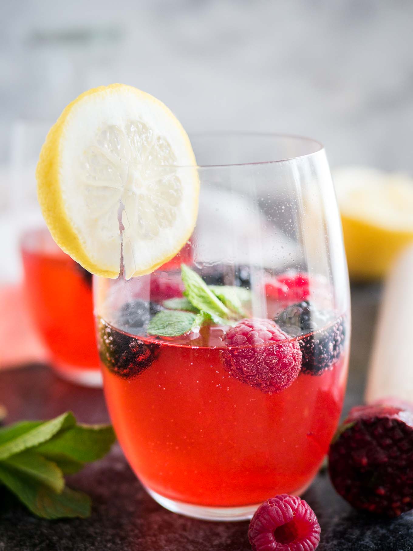 Close-up of two glasses of raspberry lemonade with raspberries and mint in them and a slice of lemon on the rim. There\'s a muddler, mint and half a lemon next to it.