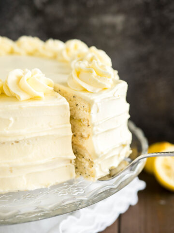 Close-up of a glass serving platter with a lemon poppy seed cake on a wooden table with a white tablecloth. A cake server is lifting out a slice.