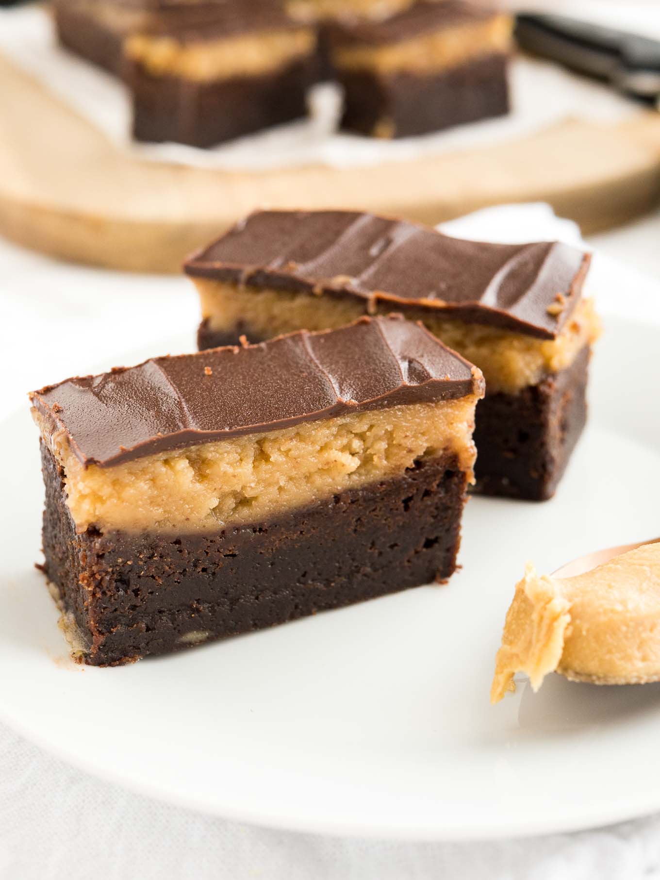 Peanut butter brownie bars on a white plate with a bronze spoon of peanut butter. A wooden cutting board with more bars is in the background.