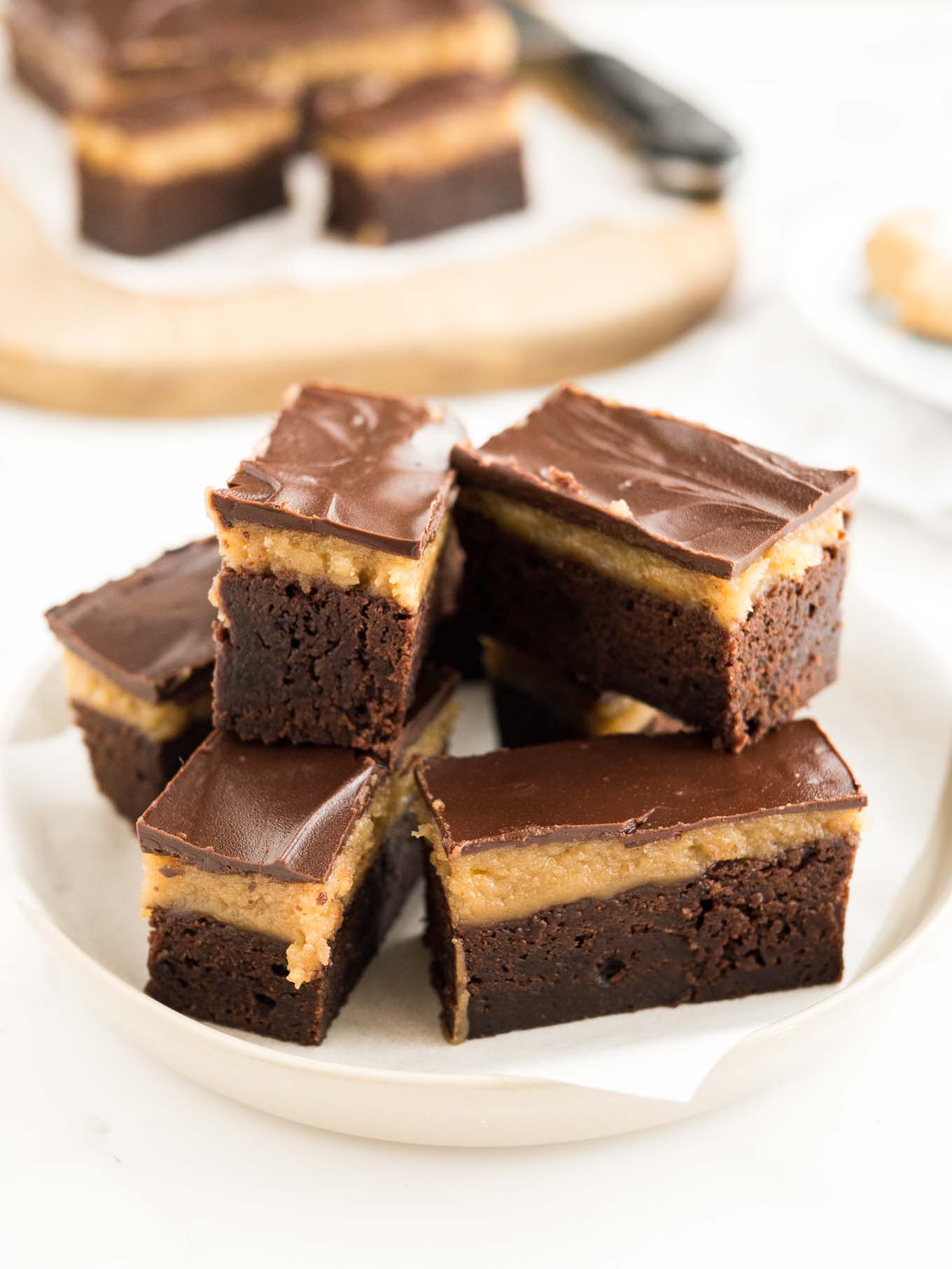 A grey plate of peanut butter brownie bars in front of a wooden cutting board with more bars on them.