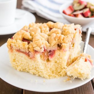 A piece of rhubarb coffee cake topped with streusel sitting on a white plate. A fork has taken out a piece of the cake. In the background, there's a cup of coffee and a small white bowl of rhubarb.