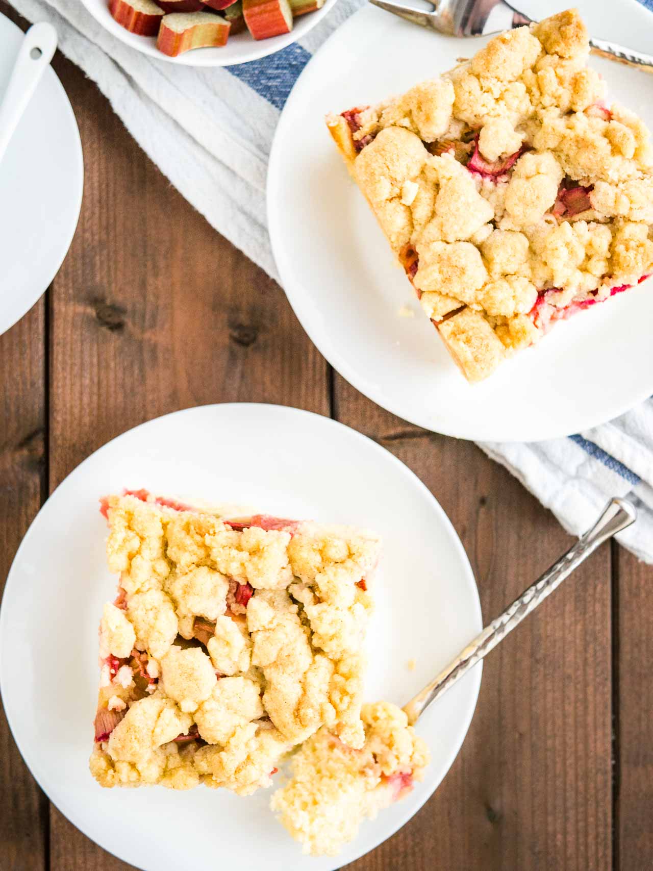 Top-down shote of a piece of rhubarb coffee cake topped with streusel sitting on a white plate. A fork has taken out a piece of the cake. Next to it, there\'s another piece of cake on a plate, a small bowl of rhubarb and a cup of coffee.