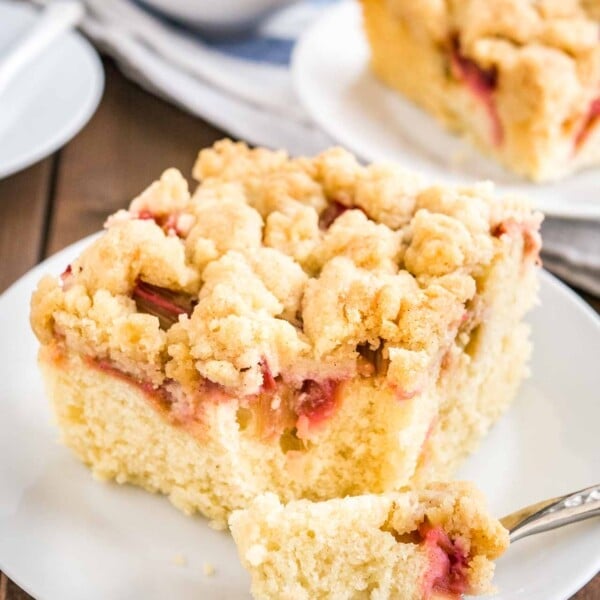 Close-up of a piece of rhubarb coffee cake topped with streusel sitting on a white plate. A fork has taken out a piece of the cake. In the background, there's a cup of coffee and another piece of cake on a plate.