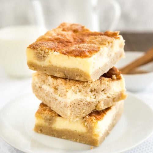 A stack of 3 snickerdoodle cheesecake bars on a white plate on a white tablecloth. There's a white bowl with cinnamon and a measuring spoon as well as a glass of milk in the background.