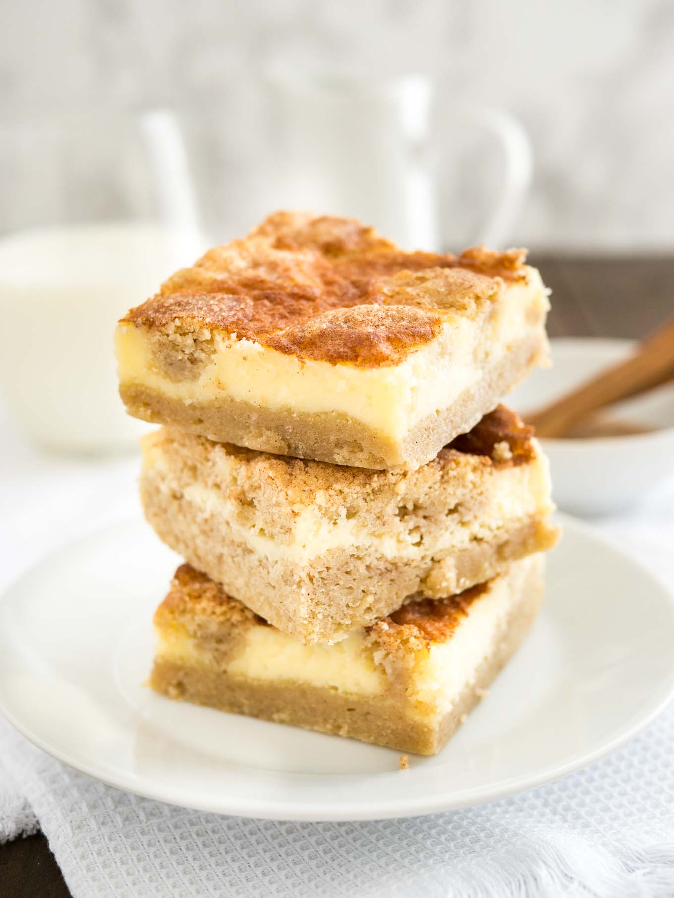 A stack of 3 snickerdoodle cheesecake bars on a white plate on a white tablecloth. There\'s a white bowl with cinnamon and a measuring spoon as well as a glass of milk in the background.