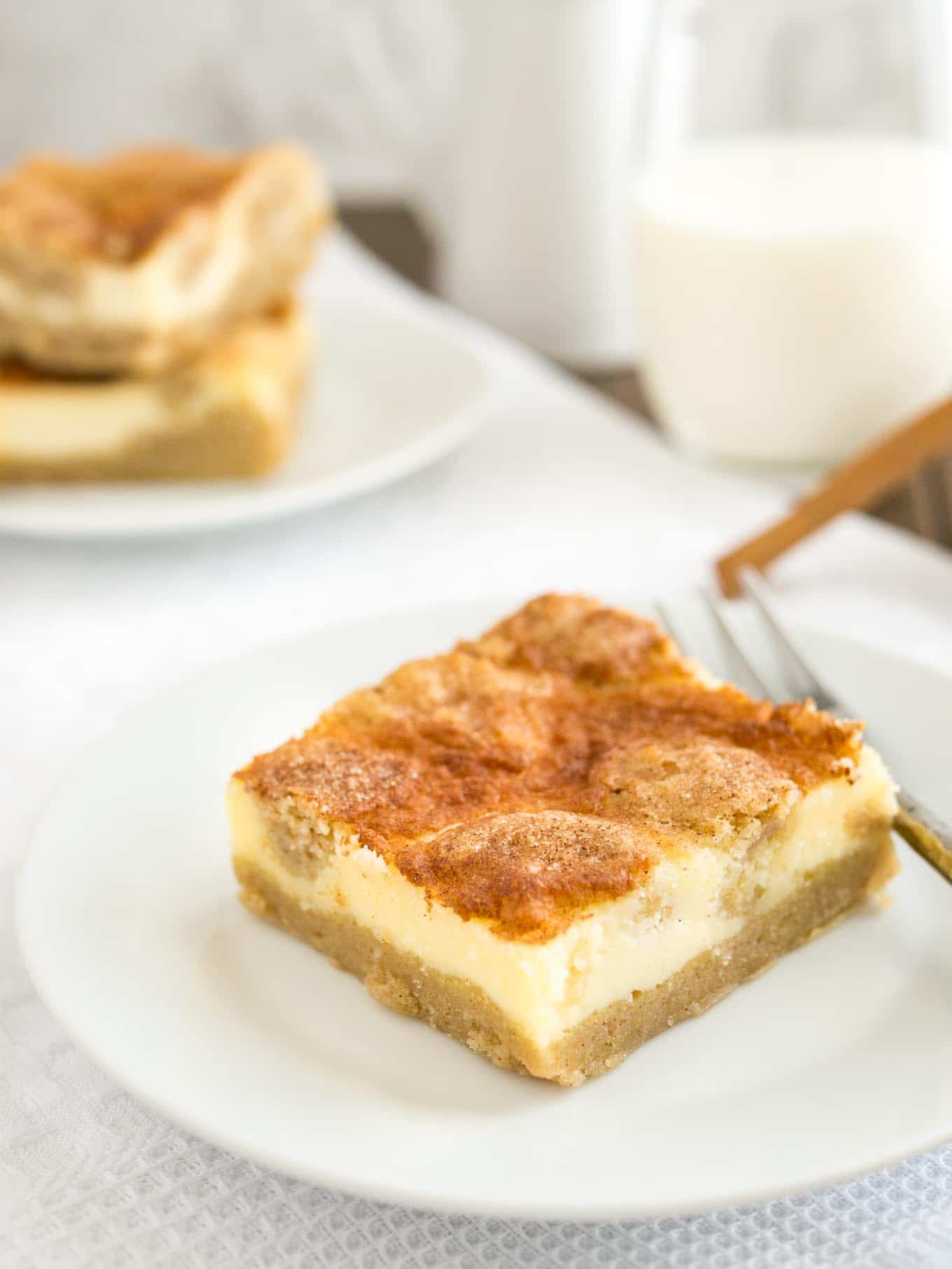 A white plate with a snickerdoodle cheesecake bar and a fork on it on a white tablecloth. There\'s a glass of milk and another plate with two cheesecake bars in the background.