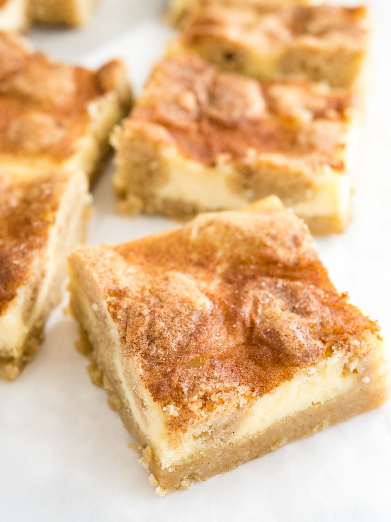 Close-up of some snickerdoodle cheesecake bars sitting on parchment paper. The frontmost is turned 45 degrees.