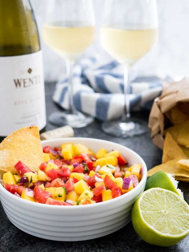 A white bowl containing mango tomato salsa with a chip in it on a dark surface next to two limes, a bag of nachos and a white and blue dish towel. There is a bottle and two glasses of white wine in the background.