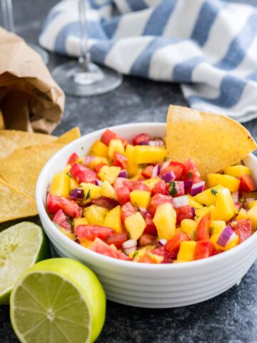 A white bowl containing mango tomato salsa with a chip in it on a dark surface next to two limes, a bag of nachos and a white and blue dish towel.