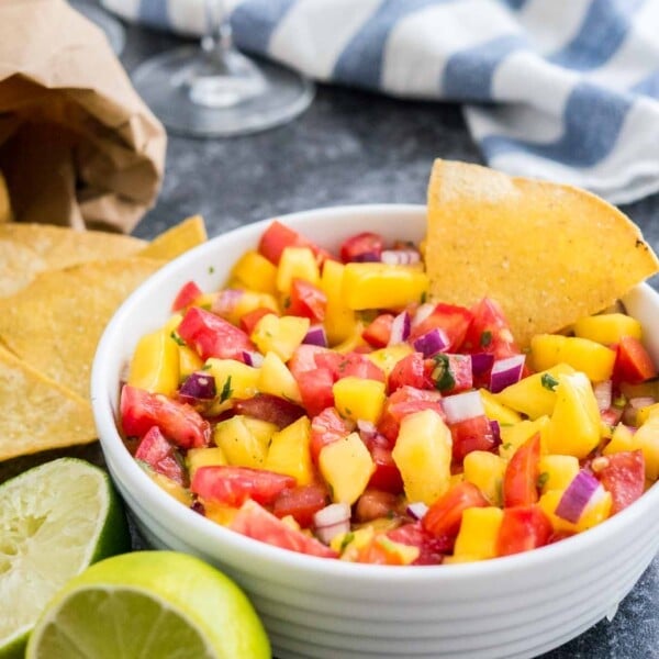 A white bowl containing mango tomato salsa with a chip in it on a dark surface next to two limes, a bag of nachos and a white and blue dish towel.