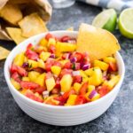 A white bowl containing mango tomato salsa with a chip in it on a dark surface next to two limes, a bag of nachos and a white and blue dish towel.