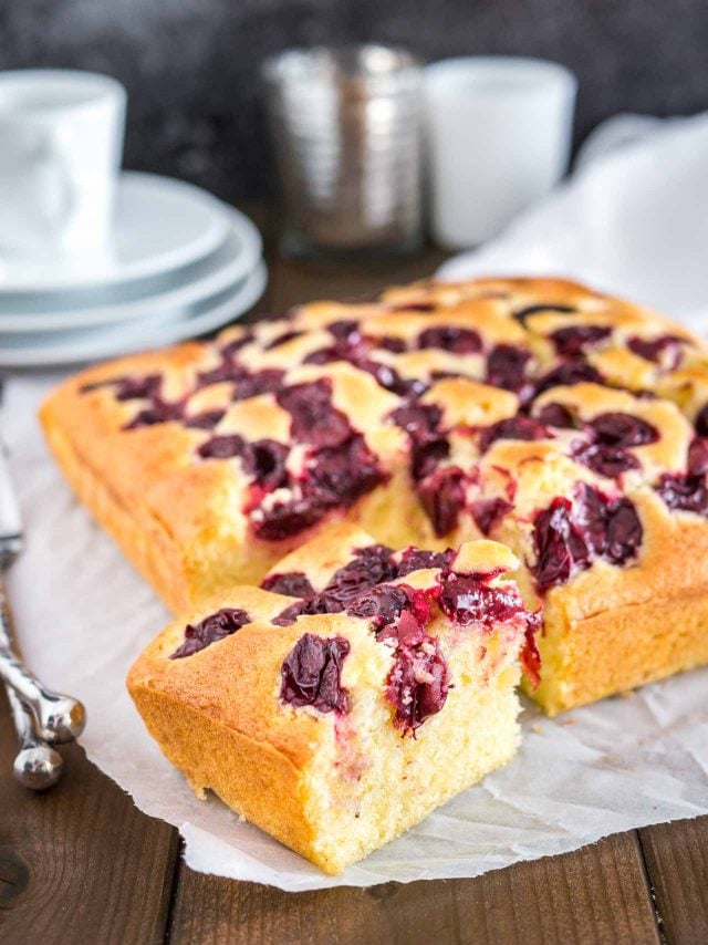 A cherry cake with one piece cut out in front of it on parchment paper. There\'s a stack of white plates with a cup on top in the background.