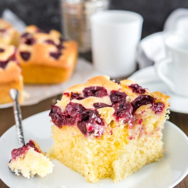 A piece of cherry cake on a white plate with a piece taken out of it by a spoon. The rest of the cake is in the background as well as a white cup.