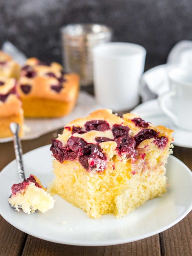 A piece of cherry cake on a white plate with a piece taken out of it by a spoon. The rest of the cake is in the background as well as a white cup.