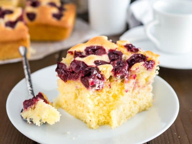 A piece of cherry cake on a white plate with a piece taken out of it by a spoon. The rest of the cake is in the background as well as a white cup.