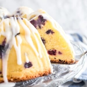 A lemon blueberry bundt cake on a glass serving platter with lemon glaze. A slice has been removed and a cake server is wedged under the cake. There's a white and blue dishtowel in the background.