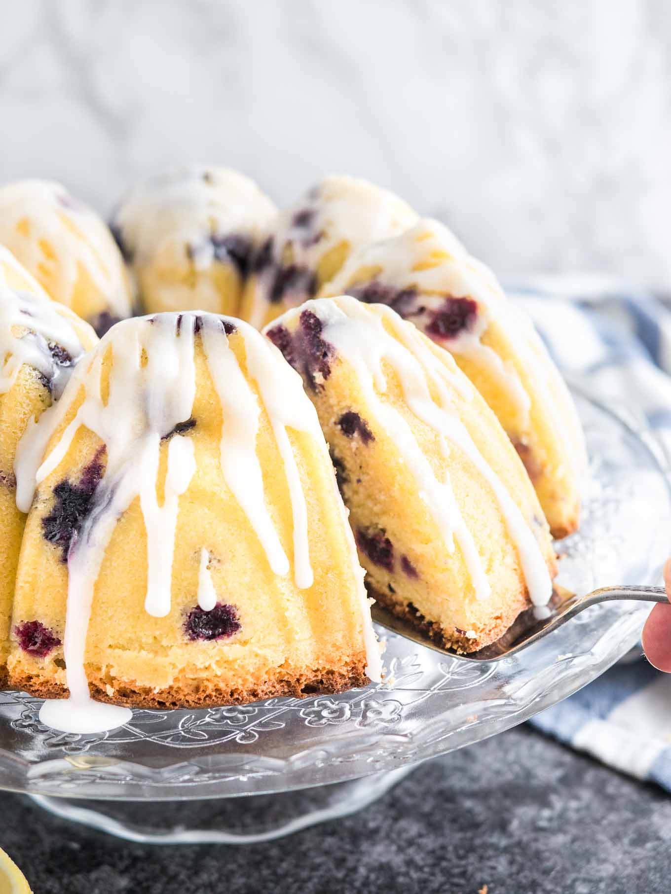 A lemon blueberry bundt cake on a glass serving platter with lemon glaze. A cake server is lifting out a slice. There\'s a white and blue dishtowel in the background.