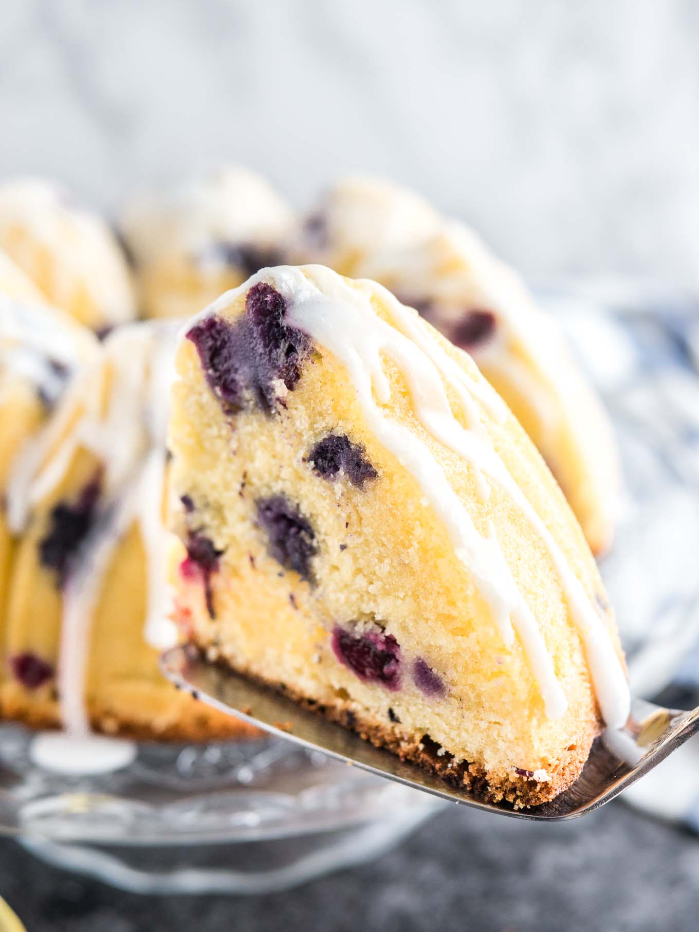 A slice of lemon blueberry bundt cake on a cake server in the foreground and the rest of the cake on a glass serving platter in the background.