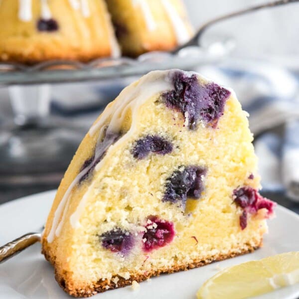 A slice of lemon blueberry bundt cake on a white plate with a fork and a wedge of lemon. The rest of the cake is on a glass serving platter in the background next to a white and blued dishtowel.