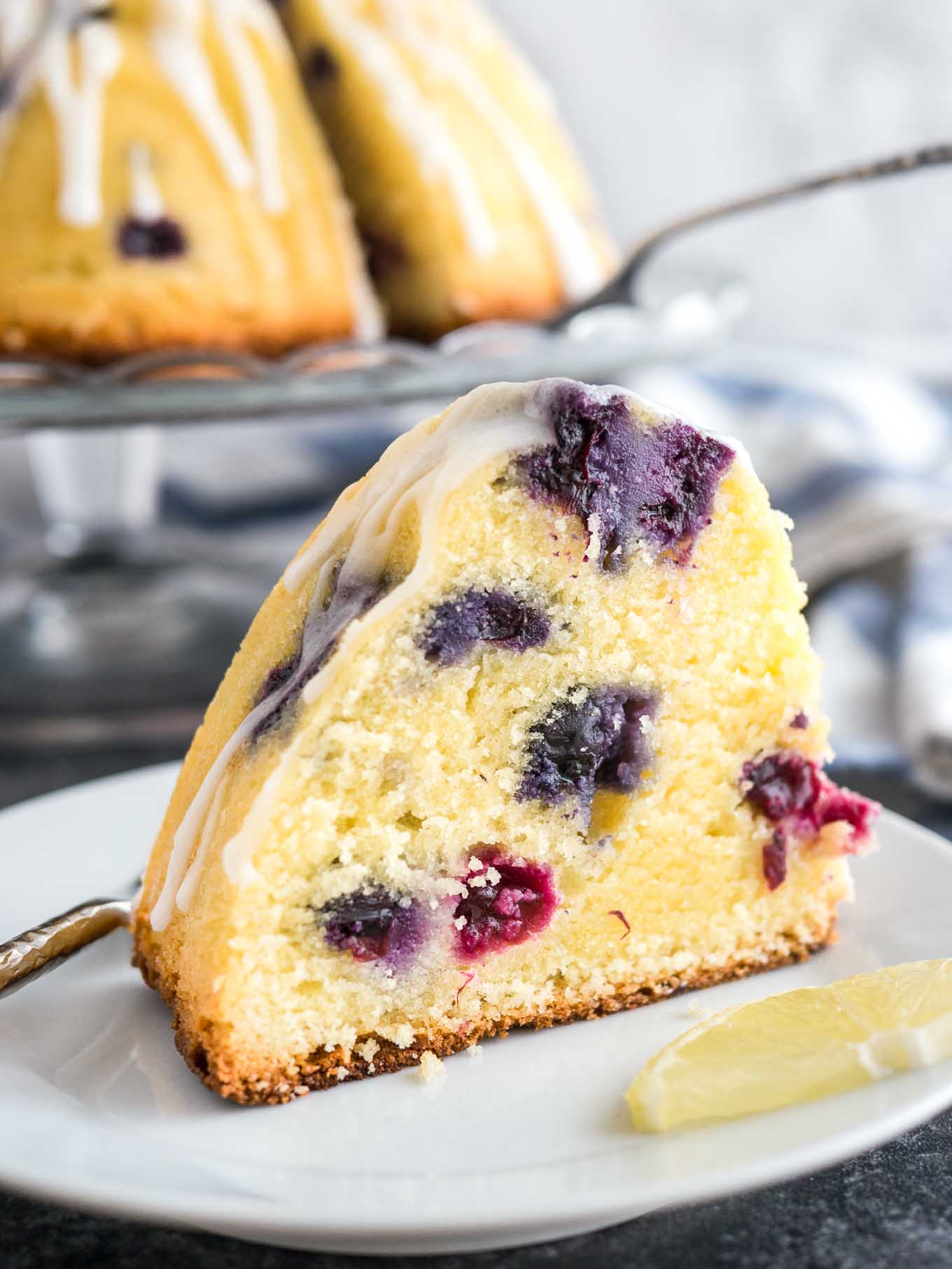 A slice of lemon blueberry bundt cake on a white plate with a fork and a wedge of lemon. The rest of the cake is on a glass serving platter in the background next to a white and blued dishtowel.