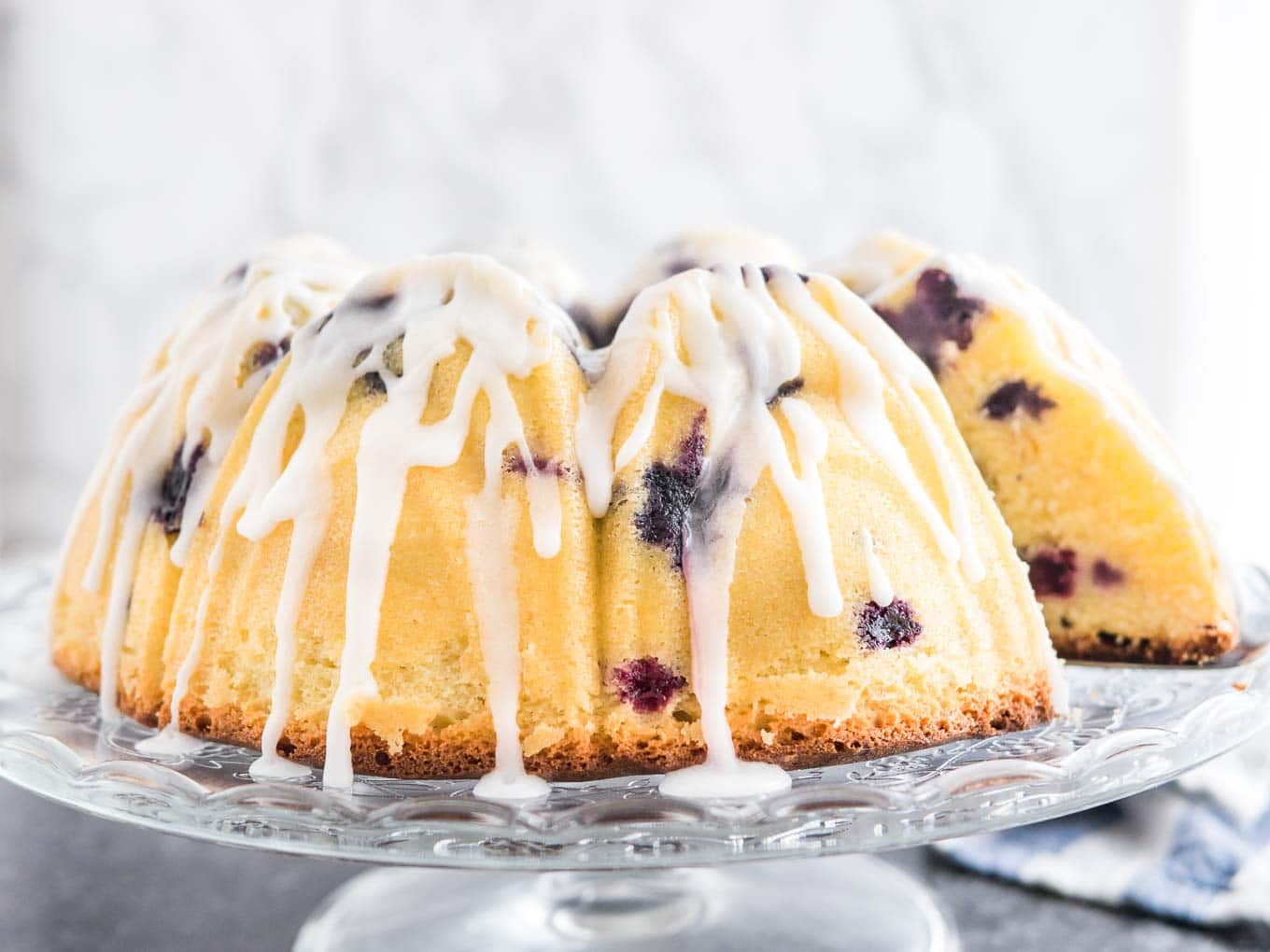 A lemon blueberry bundt cake on a glass serving platter with lemon glaze. A slice has been removed. There\'s a white and blue dishtowel in the background.
