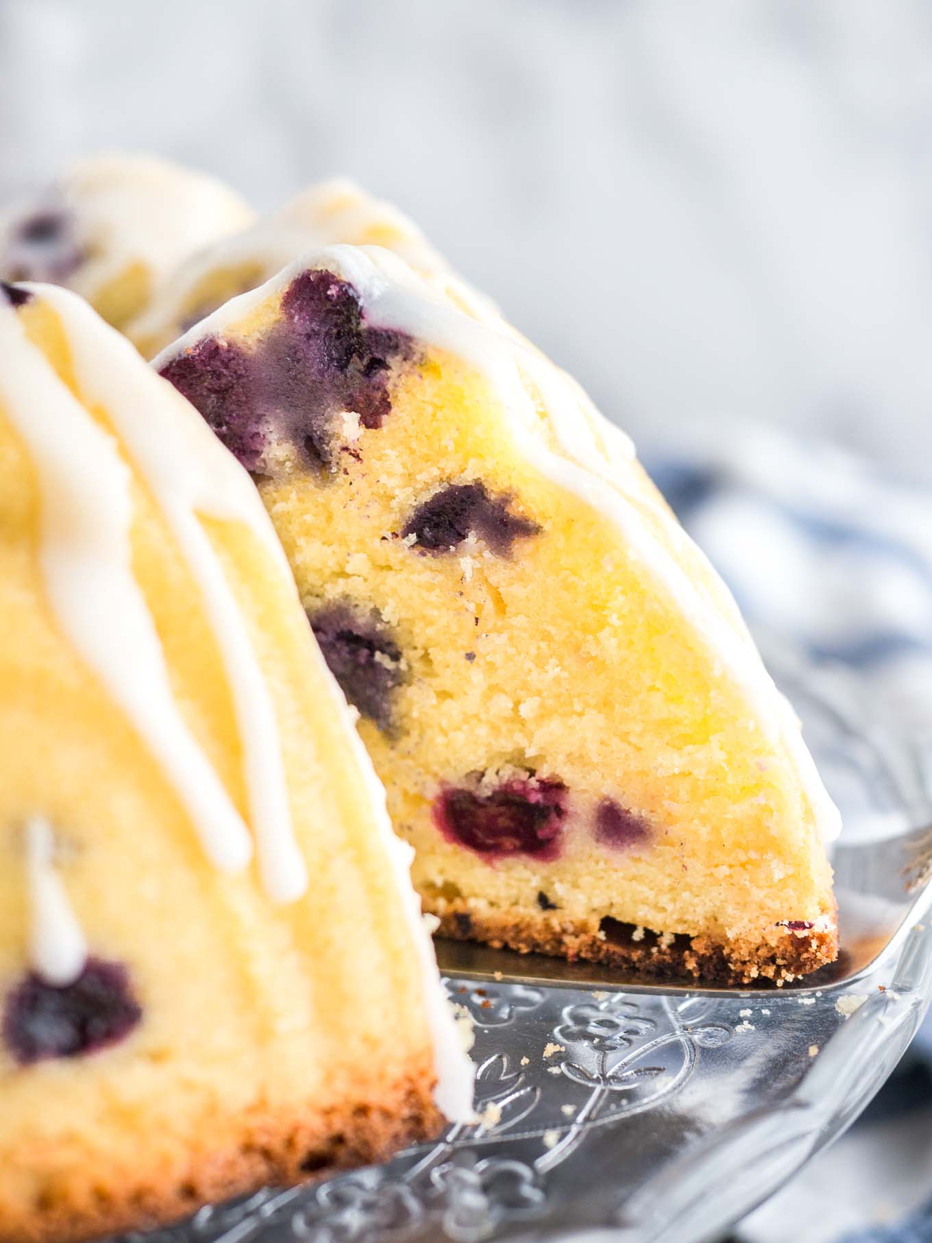 Close-up of a lemon blueberry bundt cake on a glass serving platter with lemon glaze. A slice has been removed and a cake server is wedged under the cake. There\'s a white and blue dishtowel in the background.