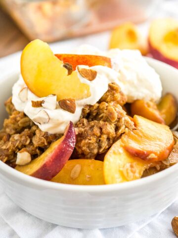 A white bowl of peach crisp with almonds and whipped cream on a white dishtowel with peach wedges in the back.