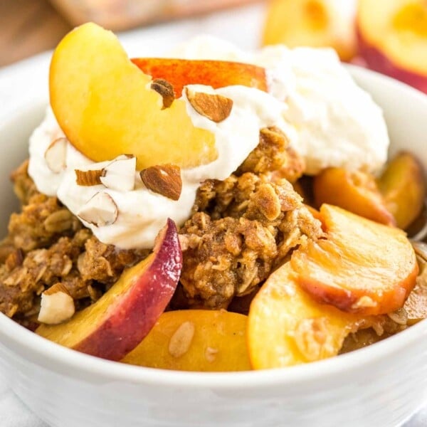 A white bowl of peach crisp with almonds and whipped cream on a white dishtowel with peach wedges in the back.
