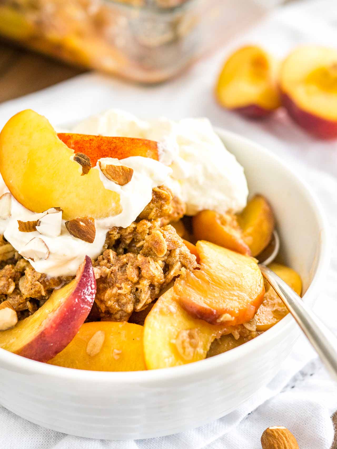 A white bowl of peach crisp with almonds and whipped cream on a white dishtowel with peach wedges in the back. A spoon is digging into the crisp.