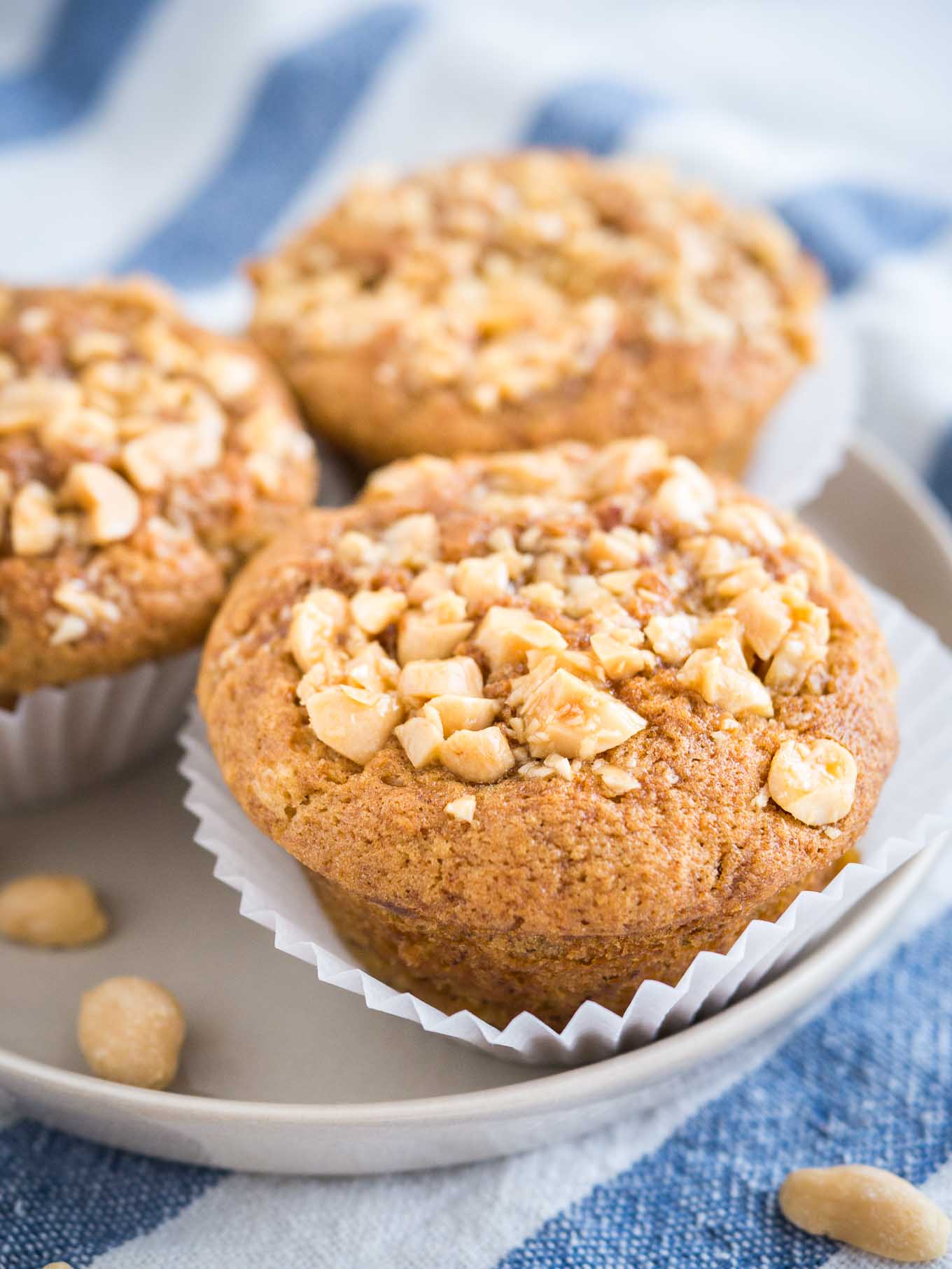 3 peanut butter banana muffins, topped with peanuts with their liners opened on a grey plate on a white and blue dishtowel.