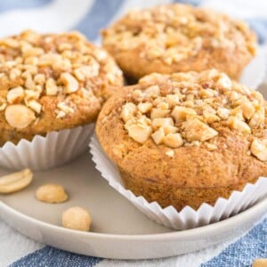 3 peanut butter banana muffins, topped with peanuts with their liners opened on a grey plate on a white and blue dishtowel.