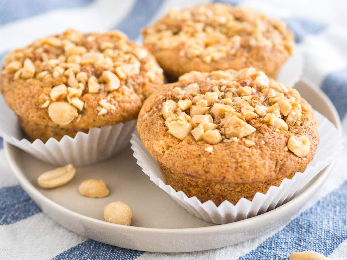 3 peanut butter banana muffins, topped with peanuts with their liners opened on a grey plate on a white and blue dishtowel.