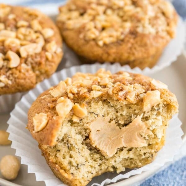 Close-up of 3 peanut butter banana muffins, topped with peanuts with their liners opened on a grey plate on a white and blue dishtowel. A big bite has been taken out of the frontmost muffin revealing a peanut butter core.