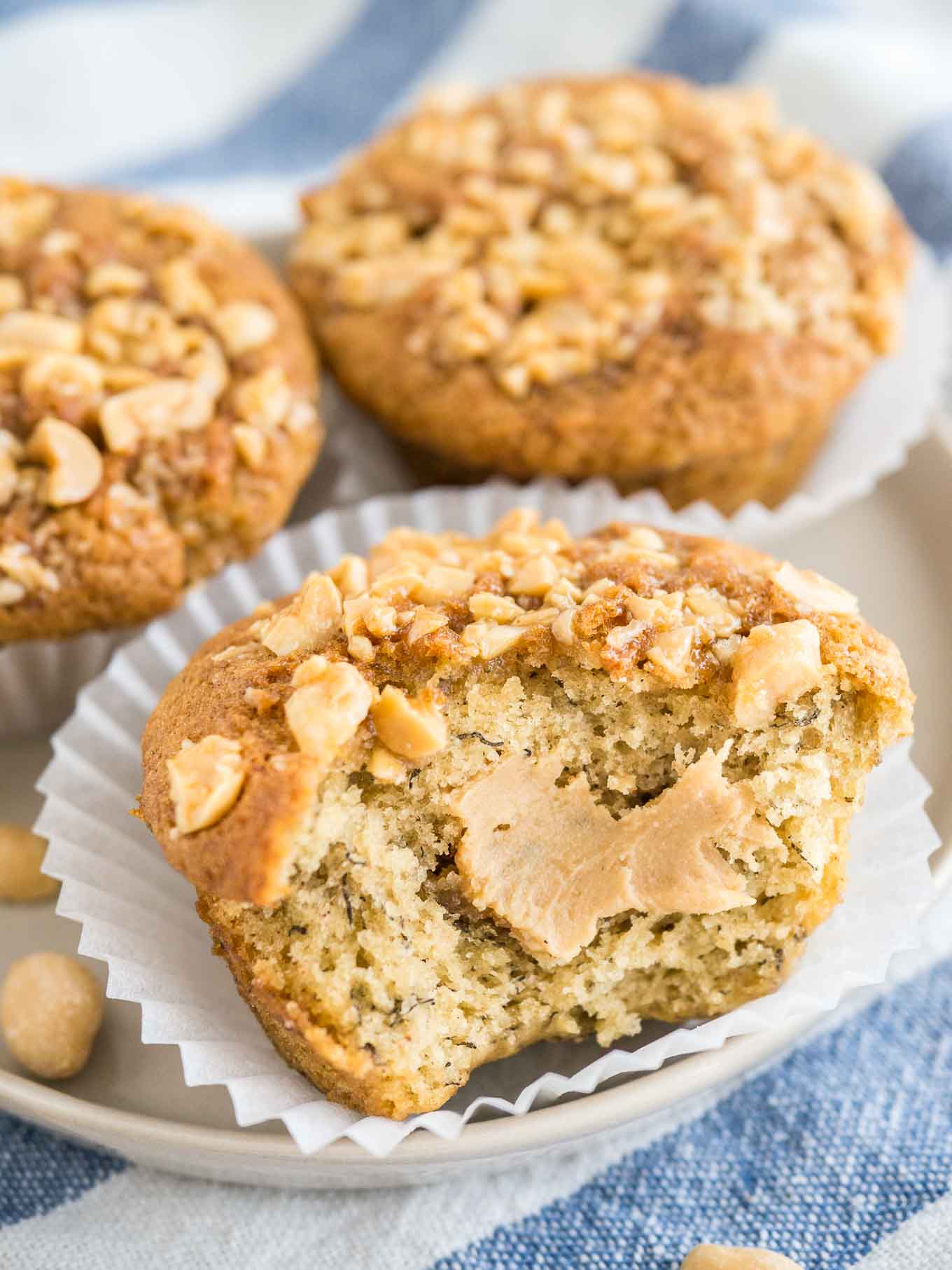 Close-up of 3 peanut butter banana muffins, topped with peanuts with their liners opened on a grey plate on a white and blue dishtowel. A big bite has been taken out of the frontmost muffin revealing a peanut butter core.