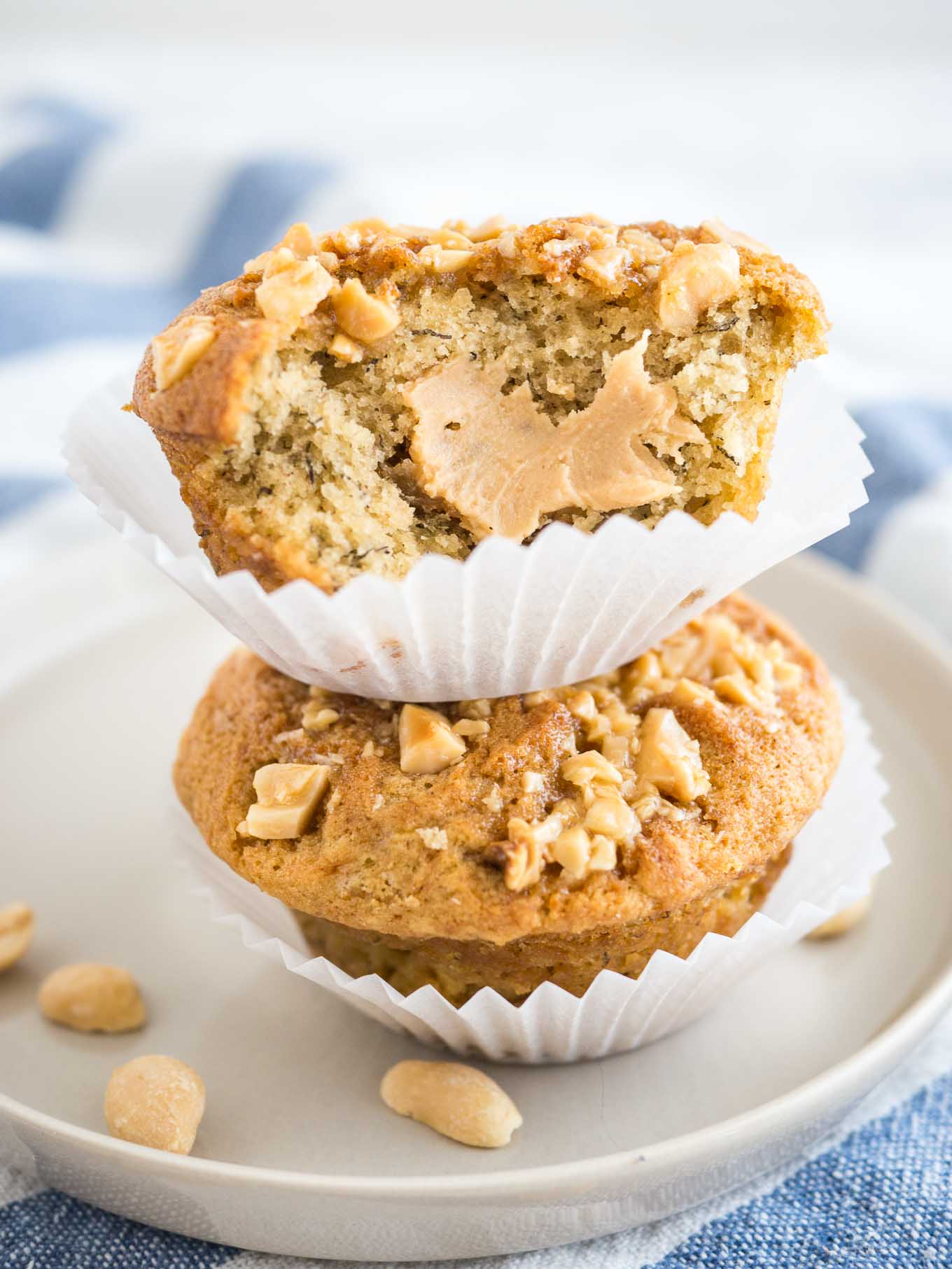 2 peanut butter banana muffins, topped with peanuts with their liners opened stacked on top of each other on a grey plate on a white and blue dishtowel. A big bite has been taken out of the top muffin revealing a peanut butter core.