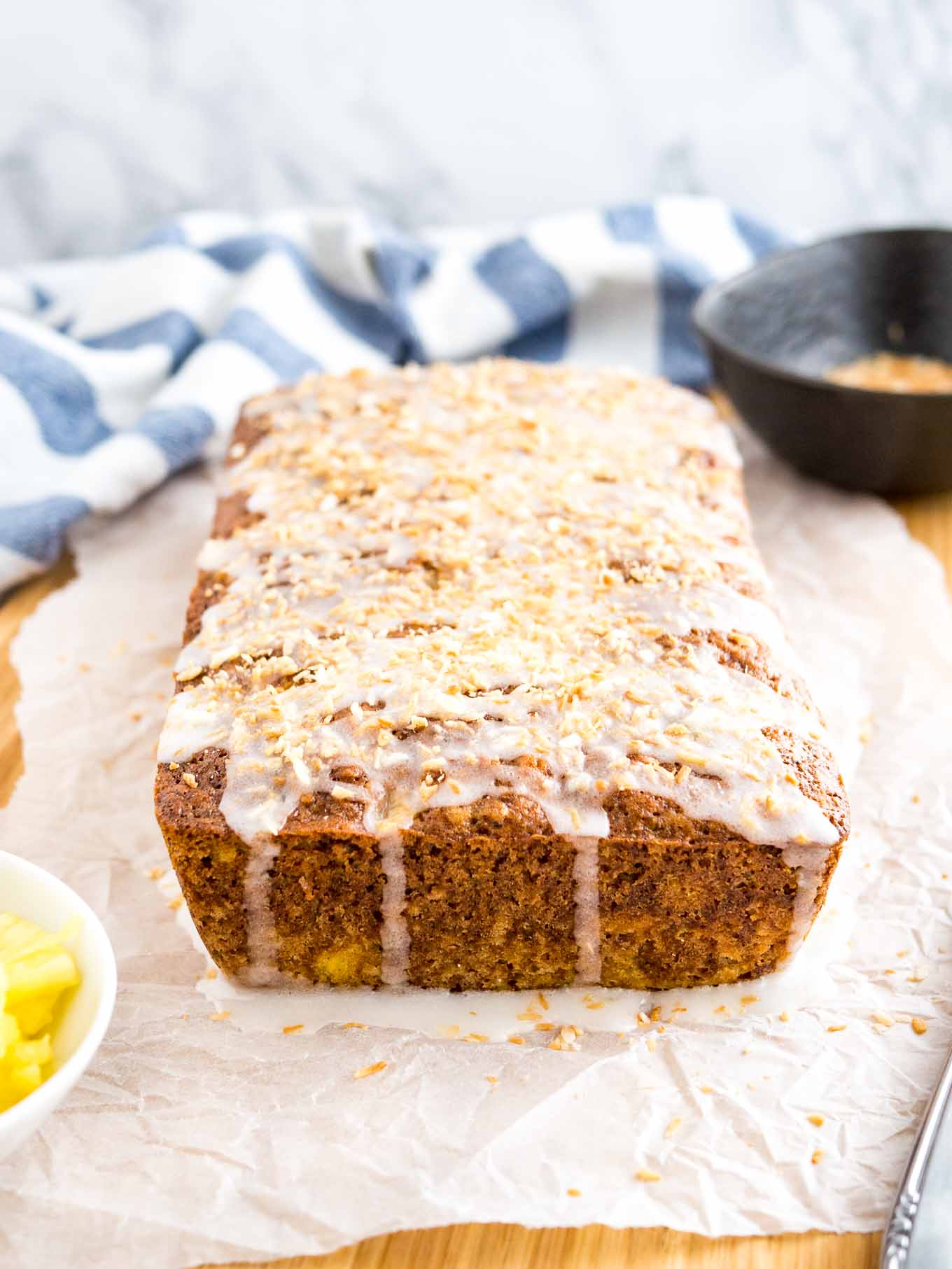 A loaf of pineapple zucchini bread on a wooden cutting board lined with parchment paper. There\'s a small white bowl of pineapple in front of it, and a small black bowl of coconut as well as a white and blue dishtowel in the background.