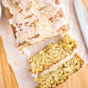 Top-down shot of a loaf of pineapple zucchini bread on a wooden cutting board lined with parchment paper. Two slices have been cut off and are lying in front of it. There's a knife, a small white bowl of pineapple and a zucchini.
