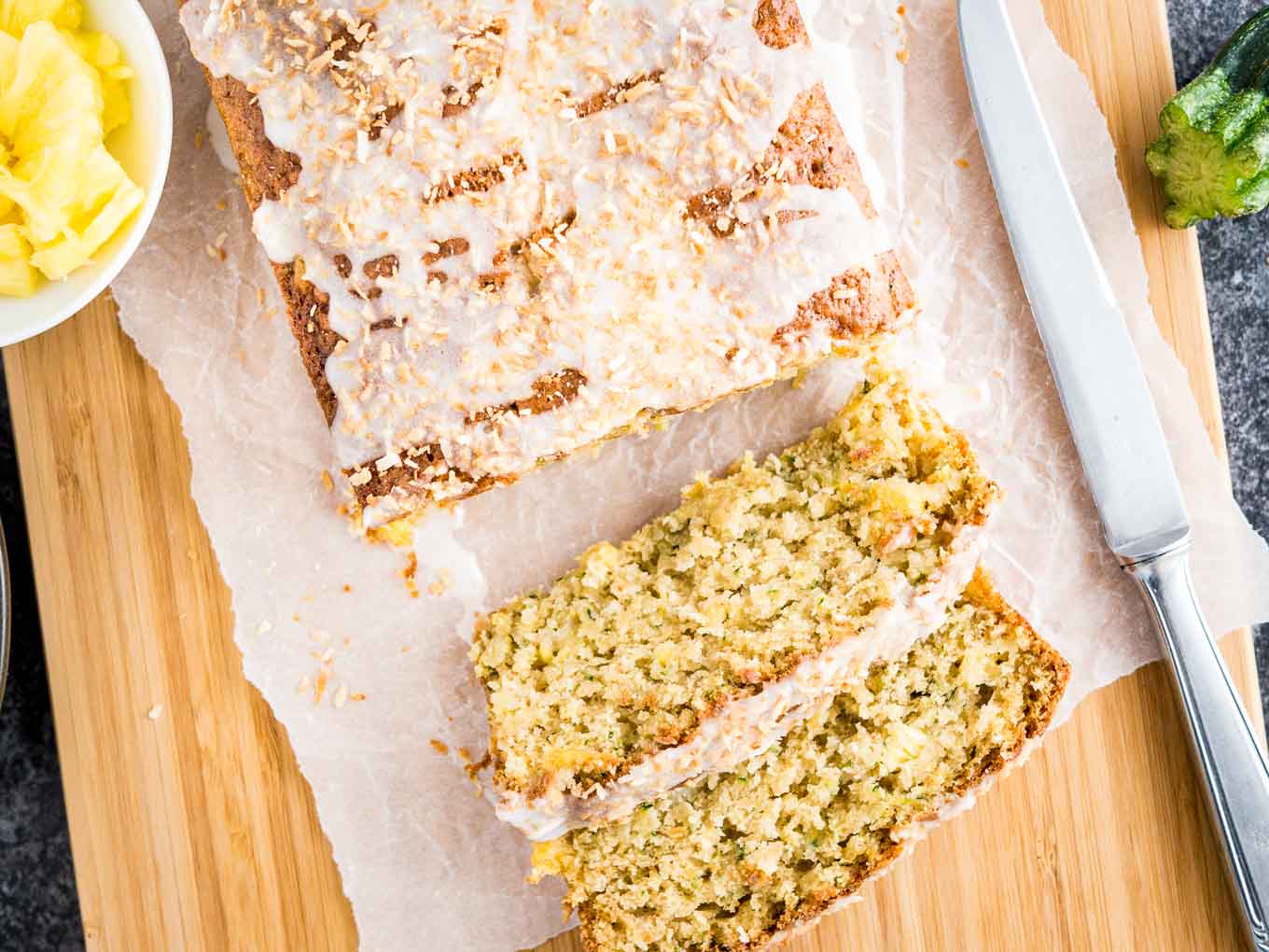 Top-down shot of a loaf of pineapple zucchini bread on a wooden cutting board lined with parchment paper. Two slices have been cut off and are lying in front of it. There\'s a knife, a small white bowl of pineapple and a zucchini.