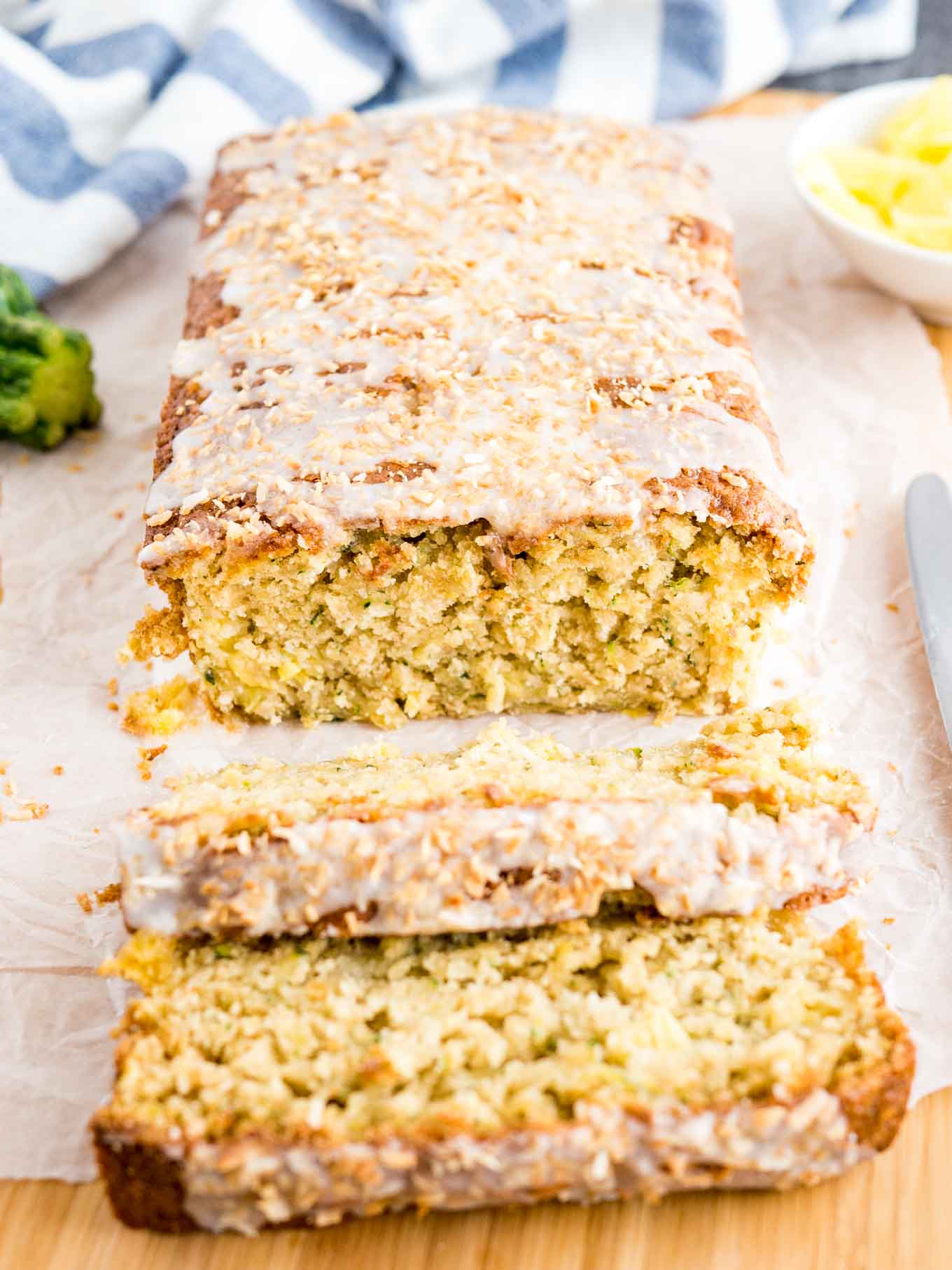 Top-down shot of a loaf of pineapple zucchini bread on a wooden cutting board lined with parchment paper. Two slices have been cut off and are lying in front of it. There\'s a knife, a small white bowl of pineapple and a zucchini.