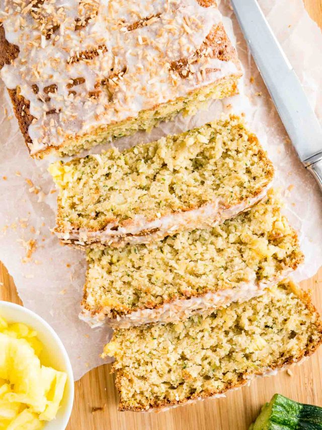 Top-down shot of a loaf of pineapple zucchini bread on a wooden cutting board lined with parchment paper. Three slices have been cut off and are lying in front of it. There\'s a knife, a small white bowl of pineapple and a zucchini.