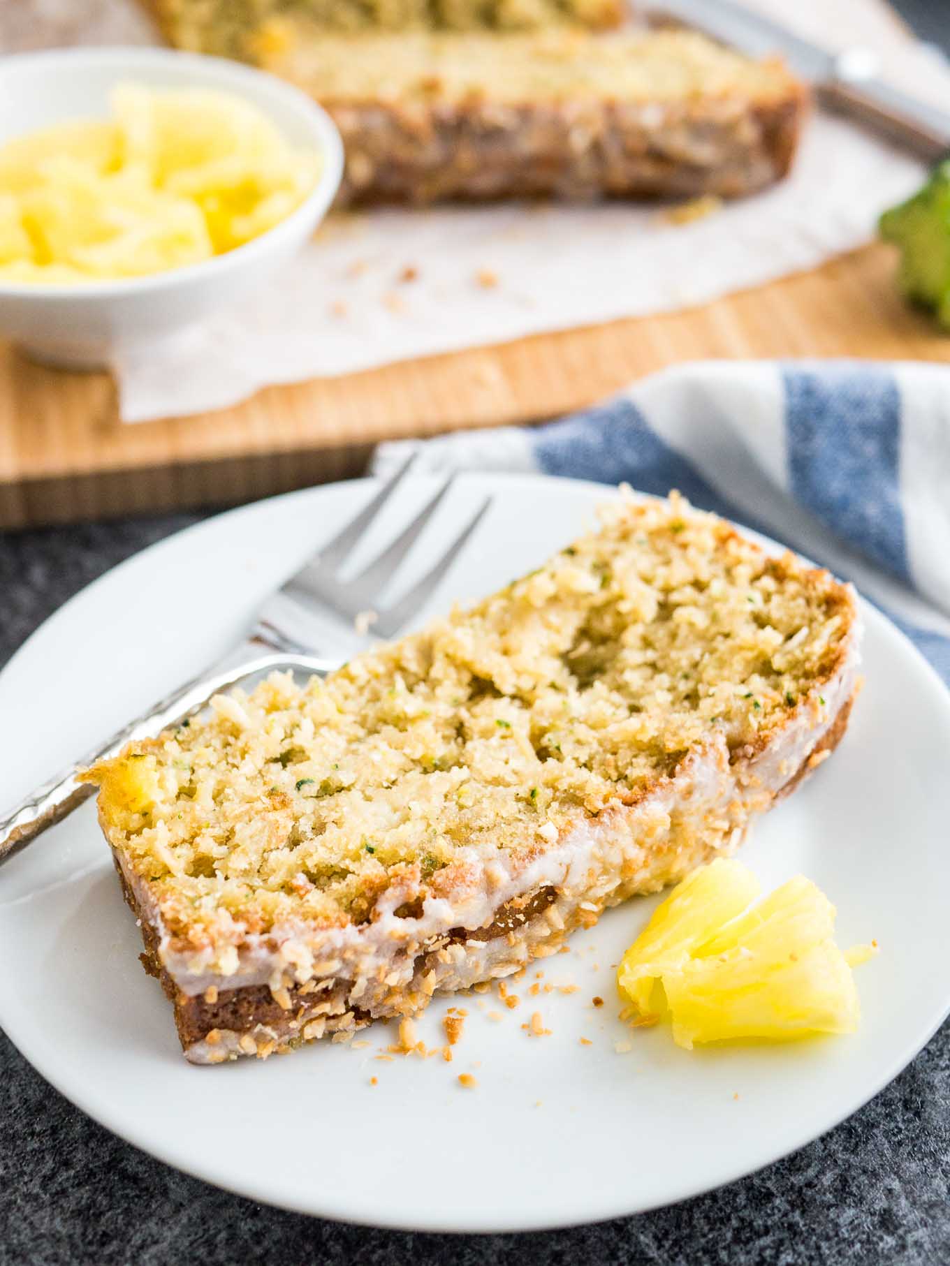 A white plate with a slice of zucchini pineapple bread with some pineapple and a fork. The rest of the bread is in the background on a wooden cutting board.