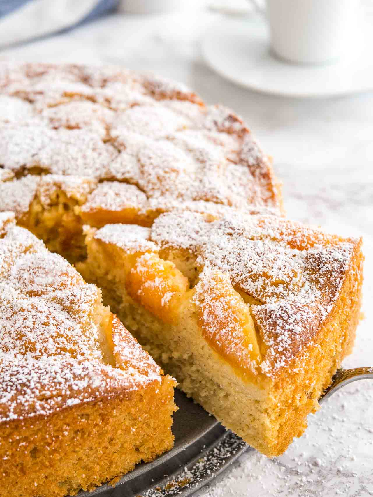 Close-up of an apricot cake on a springform bottom. A cake server is lifting out a piece and there are white coffee cups in the background.