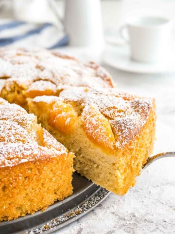 An apricot cake on a springform bottom. A cake server is lifting out a piece and there are white coffee cups in the background.