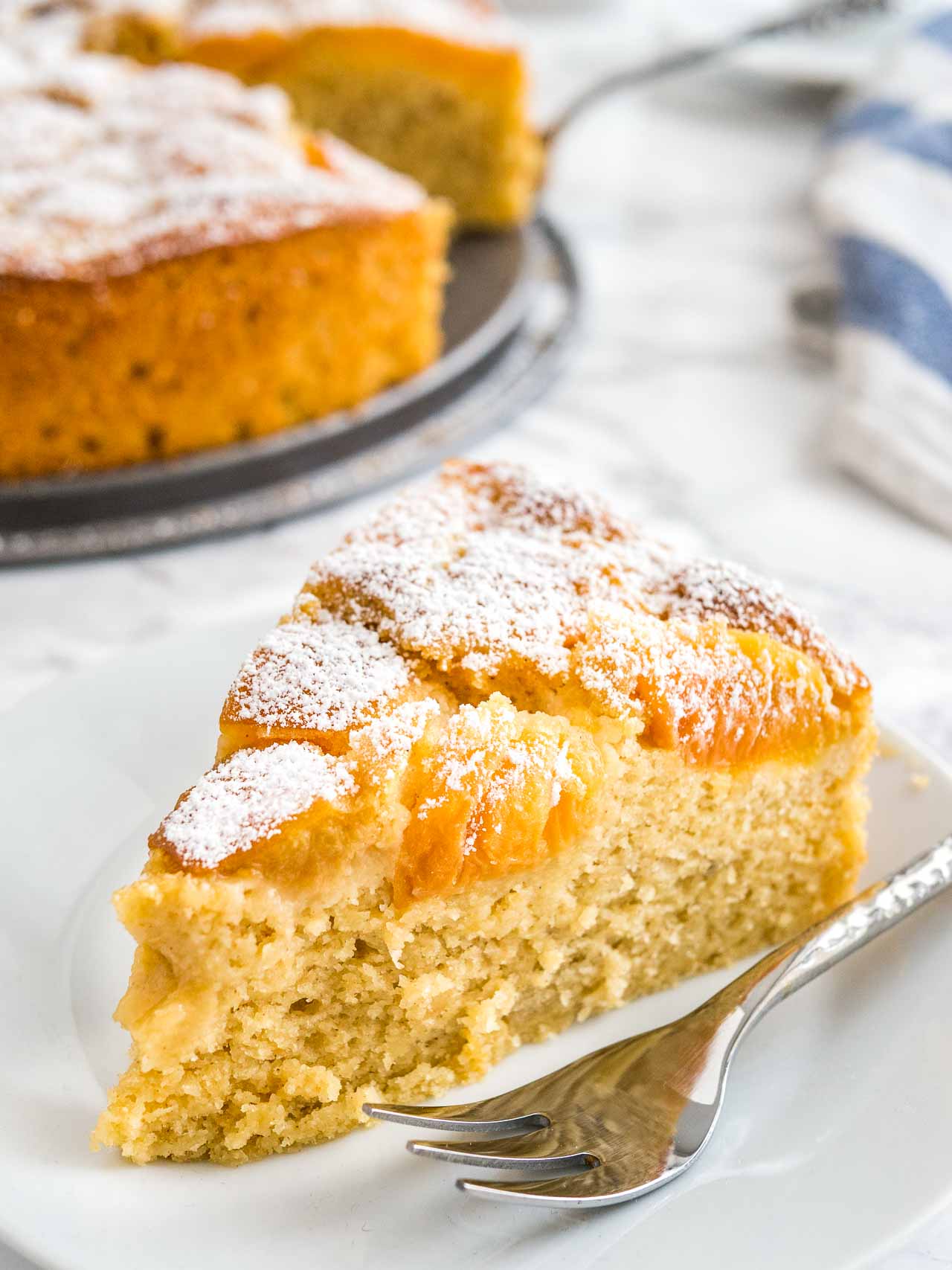 Close-up of a slice of apricot cake on a white plate with a fork. The rest of the cake is in the background next to a white and blue dishtowel.