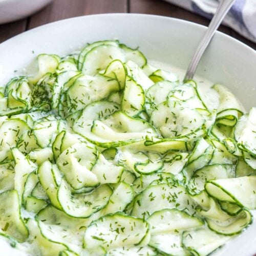 German Cucumber salad in a white salad bowl topped with dill on a wooden table with a spoon in it