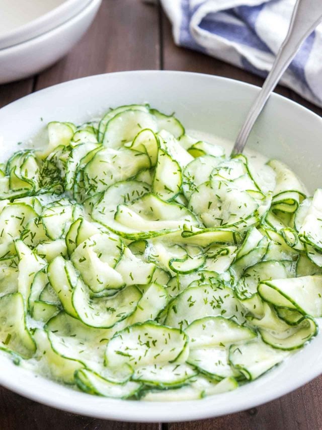 German Cucumber salad in a white salad bowl topped with dill on a wooden table with a spoon in it
